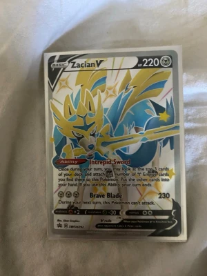 Pokémonkort Zacian V SWSH292 - Säljer ett Pokémonkort Zacian V SWSH292 med holografisk yta. Kortet har färger i blått, gult och silver och är tillverkat i laminerad papp. Perfekt för samlare eller Pokémon-fans som vill utöka sin kortsamling.