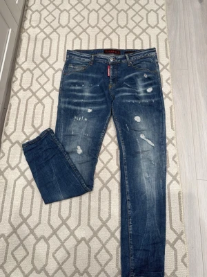 Blå slitna jeans från Icon 2 - Säljer ett par blå jeans från Icon 2 med coola slitningar och distressed detaljer. Jeansen har klassisk femficksdesign, normal midja och raka ben. Märket syns tydligt på bakfickan och vid gylfen. Perfekta för en trendig streetstyle-look.