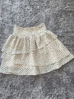 Beige vit prickig volangkjol - Söt kort kjol i beige/vit aktigtmed svarta prickar och volangdetaljer. Kjolen har en smockad midja som ger en bekväm passform och är tillverkad i ett lätt, luftigt material. 