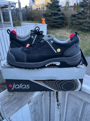 Jalas svarta sneakers med röda detaljer - Snygga svarta sneakers från Jalas med robust sula och röda detaljer vid hälen. Skorna har meshpaneler för ventilation, grå insida och reflekterande detaljer. Tillverkade i slitstarkt material och designade för både komfort och stil.    Orginal pris 3500kr