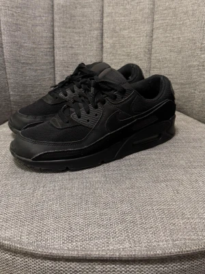 Nike Air Max 90 helsvarta sneakers - Snygga helsvarta Nike Air Max 90 sneakers med klassisk siluett. Ovandel i mesh och läder för både stil och komfort. Tjock sula med synlig Air Max-dämpning ger en sportig vibe. Perfekta för dig som gillar en clean och tidlös look.