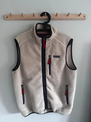 Beige fleeceväst från Patagonia - Mysig beige fleeceväst från Patagonia med hög krage och dragkedja framtill. Västen har tre fickor med dragkedja och röda detaljer samt svarta kantband. Perfekt för lager-på-lager och outdoor-vibe.