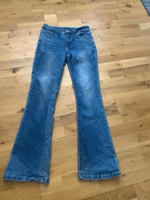 Blå utsvängda jeans med fickdetaljer - Säljer ett par blå jeans med utsvängda ben och . Jeansen har klassisk femficksdesign och bakfickor med lock och knappdetaljer. Perfekta för dig som gillar retrostil och vill sticka ut va med en cool siluett. STORLEK: M