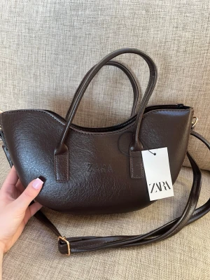 Mörkbrun handväska från Zara - Snygg mörkbrun handväska från Zara i halvmåneformad design. Perfekt accessoar för att lyfta din outfit med en trendig touch. SLUTSÅLD ❤️