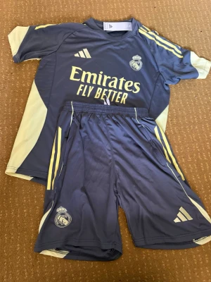 Real Madrid träningsset  - Säljer ett Real Madrid träningsset från Adidas i grått med gula detaljer. Setet består av en t-shirt och shorts med klubbmärke och Adidas-logga. Materialet är lätt och andas, perfekt för träning eller fotboll. Snygg design med gula ränder på sidorna och tryck på bröstet.