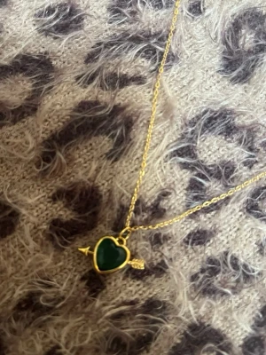 Halsband med grönt hjärthänge guld - Snyggt halsband med tunn guldfärgad kedja och ett hjärtformat hänge i grönt. Hänget har en pil genom sig vilket ger en cool och lekfull vibe. Perfekt accessoar för att lyfta din outfit med lite färg och guldtoner. Rostfri 