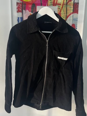 Svart manchester overshirt från The Classy Issue - Svart overshirt/vårjacka i manchester från The Classy Issue med dragkedja framtill och bröstficka med broderad logga. Klassisk krage och långa ärmar med knapp vid ärmslut. Perfekt för lager-på-lager och streetwear-stil.