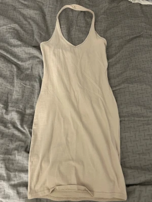 Beige halterneck klänning - En enkel och stilren beige klänning med halterneck och v-ringning. Klänningen är ärmlös och har en kroppsnära passform som framhäver siluetten. Köpt på newyorker i storlek xs, aldrig använd 