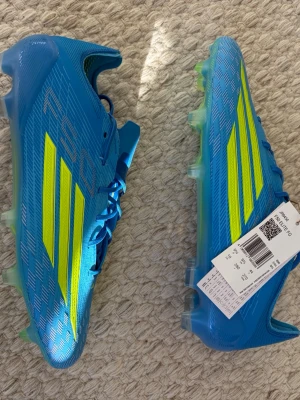 Blåa Adidas F50 fotbollsskor - Snygga blåa Adidas F50 fotbollsskor med neon-gula ränder och detaljer. Skorna har en aerodynamisk form och är tillverkade i syntetmaterial för lätt vikt och bra grepp på planen. Perfekta för dig som vill sticka ut och få max fart på fotbollen.