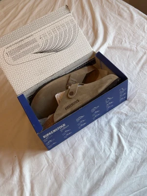Birkenstock Boston Taupe tofflor - Säljer ett par Birkenstock Boston tofflor i färgen taupe. De är tillverkade i mocka med justerbart spänne och klassisk korksula för extra komfort. Perfekta för en avslappnad och trendig stil. De är aldrig använda😁