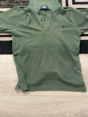 Grön piké från Ralph Lauren - Klassisk grön pikétröja från Ralph Lauren med korta ärmar och knappar vid halsen. Tröjan har den ikoniska lilla broderade loggan på bröstet och är tillverkad i mjuk bomull. Perfekt för en avslappnad och stilren look.
