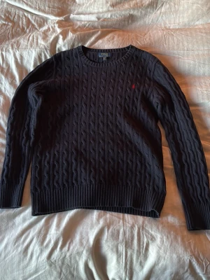 Mörkblå kabelstickad tröja Polo Ralph Lauren - Storlek XL men speciell i storlek så passar perfekt på M Kabelstickad mörkblå tröja från Polo Ralph Lauren med rund halsringning och diskret röd logga broderad på bröstet. Tröjan har långa ärmar och ribbade muddar vid ärmslut och nederkant. Perfekt för lager på lager och klassisk stil. 