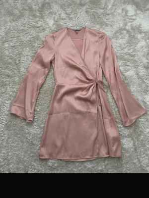 Rosa omlottklänning i satin - Snygg rosa omlottklänning i satin med långa ärmar och knyt i midjan. Klänningen har en kort längd och en elegant v-ringning. Perfekt för dig som gillar en stilren och feminin look.