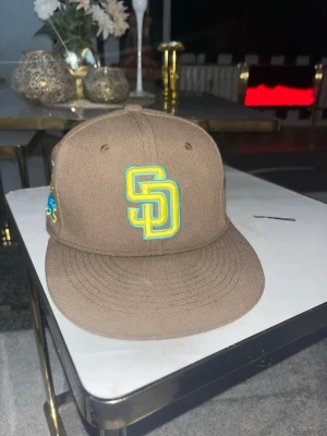 Brun San Diego Padres keps New Era - Snygg brun keps från New Era med San Diego Padres logga broderad i gult och turkost framtill. Klassisk flat peak och broderad New Era-logga på sidan. Perfekt accessoar för en sportig och trendig look.