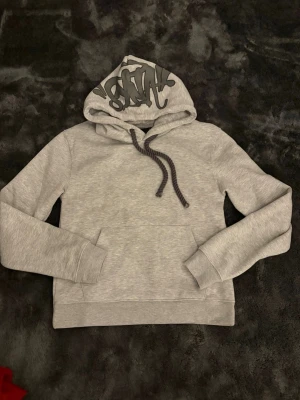 Syna World hoodie - Snygg grå hoodie från Syna world med svart tryck på huvan och matchande dragsnören. Klassisk känguruficka framtill och mjukt, borstat innerfoder för extra komfort. Perfekt för dig som gillar streetwear och vill ha en enkel men stilren look.