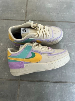 Nike Air Force 1 Shadow pastell sneakers - Nike Air Force 1 Shadow sneakers i pastellfärger med inslag av lila, mintgrönt, gult och beige. Skorna har en chunky sula, klassisk Nike Swoosh och detaljer i läder och syntet. Perfekta för dig som vill sticka ut med färg och stil.