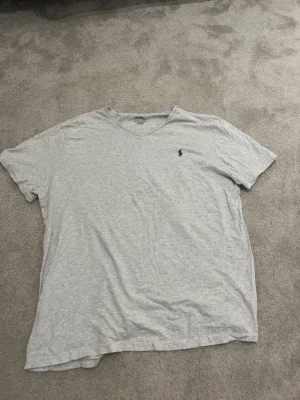 Ljusgrå v-ringad t-shirt Polo Ralph Lauren - Ljusgrå v-ringad t-shirt från Polo Ralph Lauren med klassisk liten logga broderad på bröstet. T-shirten är kortärmad och tillverkad i mjuk bomull, perfekt för en avslappnad och stilren look.
