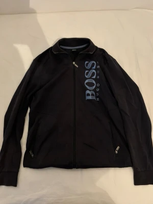 Svart tröja från Hugo Boss -  Hugo Boss tröja med dragkedja framtill och stora BOSS-tryck i ljusblått på bröstet. Tröjan har ståkrage och två fickor med dragkedja. Mjukt och bekvämt material, perfekt för chill dagar hemma.