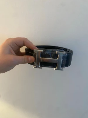 Svart skärp med H-spänne - Snyggt svart skärp  med ett stort, silverfärgat H-spänne. Skärpet har vita kontrastsömmar längs kanterna och är gjort i slätt skinn. Perfekt accessoar för att lyfta vilken outfit som helst.