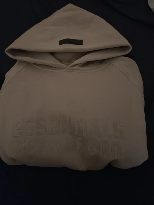 Beige Essentials Fear of God hoodie - Snygg beige hoodie från Essentials Fear of God med stor huva och diskret logga på bröstet. Hoodien har en klassisk känguruficka och ribbade muddar vid ärmar och nederkant. Perfekt för en avslappnad och trendig streetwear-look. PASSAR S OCH XS