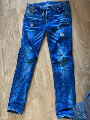 Blå distressed jeans med färgstänk - Säljer ett par blå jeans med slitningar, färgstänk och unika detaljer. Jeansen har smal passform, klassisk femficksdesign och dragkedja framtill. Materialet är denim i bomull och de har en trendig, streetinspirerad look. Storlek S och inte använt mycket alls, bara ett par gånger. Det går bra att pruta men skriv inte om du inte tänker köpa