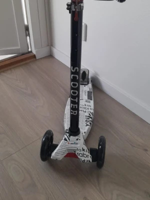 Svart och vit sparkcykel med tidningsmönster - En cool trehjulig sparkcykel i svart och vitt med tidningsinspirerat mönster på fotplattan. Styrstången är svart med texten SCOOTER i vitt. Perfekt för att cruisa runt i stan eller på gården.