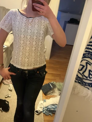 Vit spets-topp med kort ärm - Söt vit topp i spets med korta ärmar och rund svart halsringning. Tunt, blommigt spetsmönster över hela toppen och lätt genomskinlig. Svart kantband vid halsen ger en snygg kontrast. Perfekt att styla.