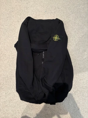 Svart hoodie från Stone Island - Svart hoodie från Stone Island med dragkedja och klassisk logotyp-patch på vänster ärm. Hoodien har en enkel och stilren design med huva och ribbade muddar. Perfekt för dig som gillar streetwear och vill ha något snyggt och bekvämt.