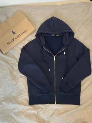 Mörkblå hoodie från Polo Ralph Lauren - Snygg mörkblå hoodie från Polo Ralph Lauren med dragkedja framtill och klassisk huva. Ikoniska loggan broderad på bröstet. Mjukt material och ribbade muddar vid ärmslut och nederkant. Perfekt för en avslappnad och stilren look.