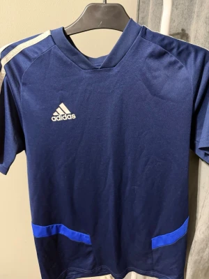 Blå Adidas träningströja ClimaCool - Mörkblå träningströja från Adidas med vita detaljer och blåa inslag vid midjan. Tillverkad i lätt och ventilerande ClimaCool-material som passar perfekt för sport och träning. Klassisk Adidas-logga på bröstet och korta ärmar.