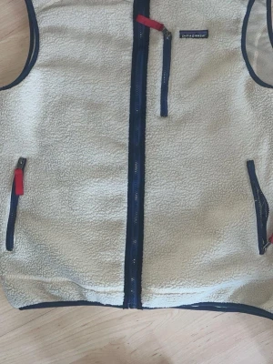 Beige fleeceväst från Patagonia - Säljer en beige fleeceväst från Patagonia med marinblå detaljer och röda dragkedjor. Västen har två sidofickor med dragkedja och en bröstficka. Perfekt för lager-på-lager och outdoor-stil.