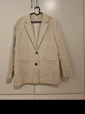 Beige kostym med kavaj och byxor - Snygg beige kostym med kavaj och matchande byxor. Kavajen har två knappar och raka slag, medan byxorna har raka ben och fickor på sidorna. Perfekt för en stilren och modern look.