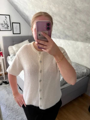 Vit stickad kofta med kort ärm - Mysig vit stickad kofta med korta ärmar och rund hals. Koftan har ribbad struktur och knäpps med pärlemorknappar framtill. Perfekt att slänga på sig över ett linne eller t-shirt för en chill look.