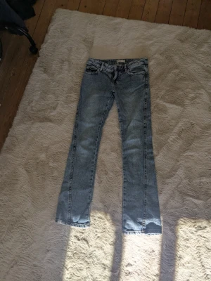 Ljusblå bootcut jeans med låg midja  - Säljer ett par ljusblå bootcut jeans med klassisk femficksdesign och dragkedjegylf. Jeansen har en lätt tvättad look och raka ben som går ut lite nertill. Perfekta för dig som gillar en avslappnad och tidlös stil.
