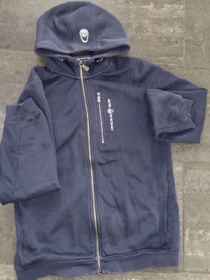 Marinblå hoodie från Sail Racing - Snygg marinblå hoodie från Sail Racing med dragkedja framtill och en bröstficka med logga. Hoodien har en broderad logotyp på huvan och är fodrad med mjuk fleece för extra komfort. 