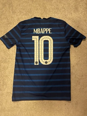 Frankrike Mbappé matchtröja blå - Säljer en blå matchtröja från Nike med Frankrikes landslagsmärke och Mbappé 10 på ryggen. Tröjan har mörkblå och ljusblå ränder, röd detalj på bröstet och korta ärmar. Perfekt för fotbollsfans som vill stötta sitt lag.