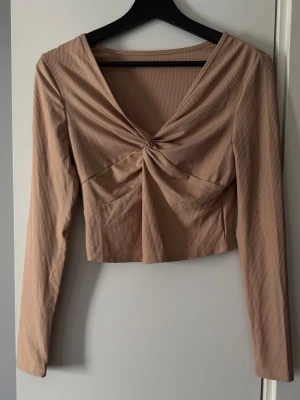 Beige croppad topp med twistad front SHEIN - Trendig beige croppad topp från SHEIN med lång ärm och twistad detalj framtill. Toppen har v-ringning och är tillverkad i ett ribbat material som ger en snygg struktur. Perfekt för dig som gillar stilrena och moderna plagg.