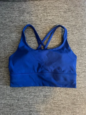 Blå sport-bh från Lululemon - Snygg blå sport-bh från Lululemon med korsade axelband i ryggen. Tillverkad i ett stretchigt och mjukt material som ger bra stöd och komfort. Perfekt för träning eller som en del av din vardagslook.