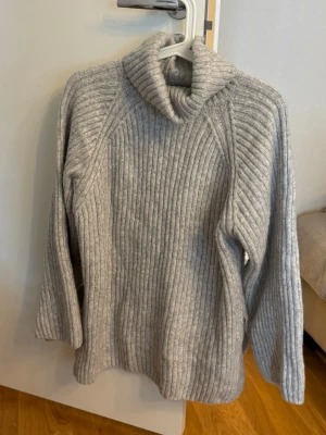 Grå ribbstickad polotröja - Mysig grå stickad polotröja med bred ribbstickning och långa ärmar. Tröjan har en hög, vikt krage och är tillverkad i ett mjukt material som passar perfekt för kyliga dagar. Enkel och stilren design som är lätt att matcha.