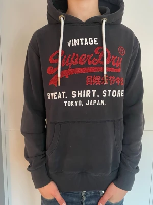 Svart Superdry hoodie med tryck - Snygg svart hoodie från Superdry med stort rött och vitt tryck på bröstet och texten 'Vintage Superdry Sweat. Shirt. Store Tokyo, Japan'. Klassisk känguruficka, huva med vita snören och mjukt material. Perfekt för en avslappnad streetwear-look.