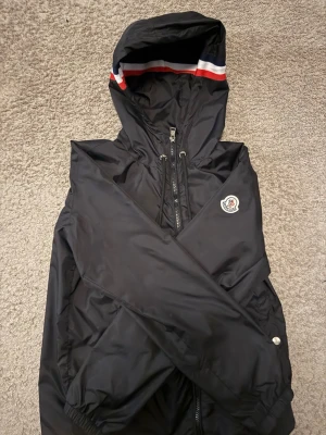 Moncler vindjacka - Snygg svart vindjacka från Moncler med huva och dragkedja framtill. Jackan har Moncler-logga på ärmen och en cool detalj med rödvit rand på huvan. Tillverkad i lätt och tåligt material, perfekt för blåsiga dagar. Nfc tagg i moncler loggan