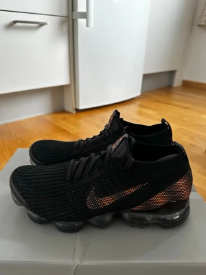 Nike Vapormax svarta sneakers - Nike Vapormax sneakers i svart med orange detaljer och stickad ovandel. Snygg, sportig design med synlig luftdämpad sula och diskret Nike-logga på sidan. Ovandelen är i mesh för extra andningsförmåga och komfort. Perfekta för en streetwear-look. Nya använda max 1-2 gånger