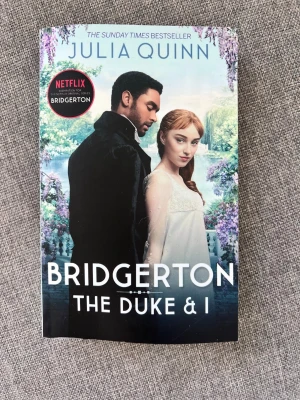 Bridgerton: The Duke & I - En romantisk och historisk roman av Julia Quinn, känd från Netflix-serien Bridgerton. Boken har ett färgstarkt omslag med två karaktärer mot en blomstrande bakgrund. Perfekt för dig som gillar kärlek, drama och 1800-talets London.