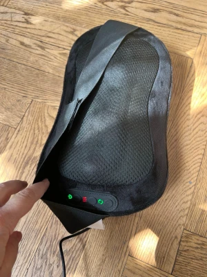 Elektrisk massagekudde svart - Elektrisk massagekudde i svart med mesh-tyg och mjukt material. Kudden har ett justerbart band och kontrollpanel med knappar för värme och massagefunktion. Perfekt för att slappna av hemma eller på kontoret. Strömkabel medföljer.