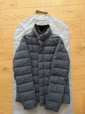 Grå dunjacka från Herno - Säljer en snygg grå dunjacka från Herno med hög krage och mjukt brunt foder på insidan av kragen. Jackan har quiltad design och stängs med både dragkedja och tryckknappar. Perfekt för kalla vinterdagar och riktigt stilren. Storlek 50, passar M-L