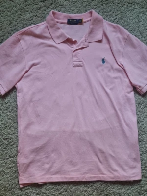 Rosa pikétröja från Polo Ralph Lauren - Klassisk rosa pikétröja från Polo Ralph Lauren med blå broderad logga på bröstet. Tröjan har krage, korta ärmar och knäppning framtill. Perfekt för en stilren och avslappnad look.