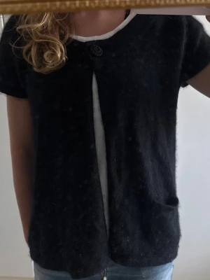 Svart kortärmad kofta med knapp och fickor - Svart kofta med korta ärmar, två fickor och en stor knapp upptill. Koftan är öppen framtill och har en avslappnad passform. Perfekt att slänga över en t-shirt för en chill look.