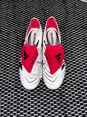 Adidas Predator Elite - Säljer helt oanvända Adidas Predator. Skorna har järn dobbar för dom är gjorda för mjukmark/gräs (SG). Dom är vita med en röd tunga, tungan har band som går över skorna där det står predator, skorna har också gummi på insida skorna där du kan skruva och få bättre grepp på bollen. Pris kan självklart diskuteras.