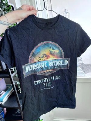 Jurassic World svart t-shirt - Svart t-shirt med färgglatt Jurassic World-tryck på bröstet och texten 'Exklusivt på bio 2 juli'. Klassisk rund hals och korta ärmar. Perfekt för dig som älskar film och coola prints.