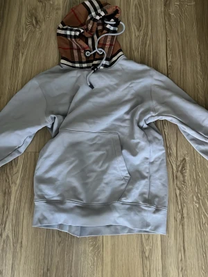 Grå hoodie med Burberry-rutig huva - Snygg grå hoodie från Burberry med klassisk rutig huva i beige, svart, vitt och rött. Hoodien har en stor magficka och dragsko i huvan. Perfekt för dig som vill ha en stilren men ändå unik look. Materialet känns mjukt och skönt mot huden.